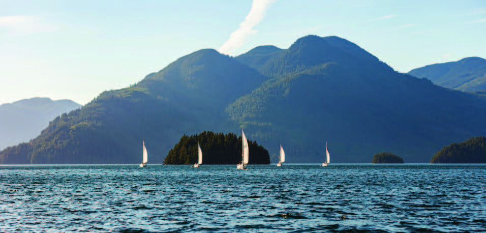 5 sailboats out at sea, forested ranges in the background.